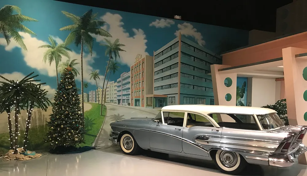 The image features a classic car parked in front of a vibrant mural depicting a tropical street scene complete with palm trees and Art Deco-style buildings