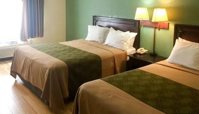 The image shows a modest hotel room with two neatly made double beds a nightstand with a phone and a window with curtains to one side