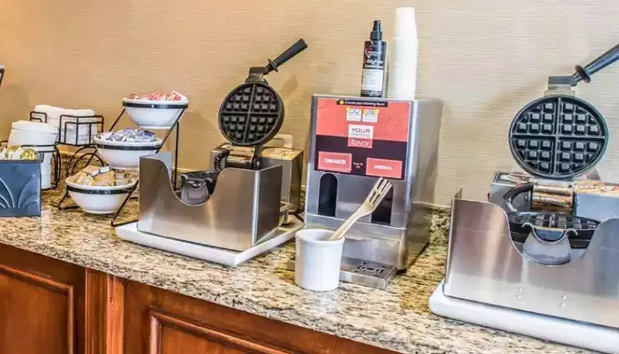 The image shows a self-service waffle station with batter dispensers and waffle irons, part of a continental breakfast setup.