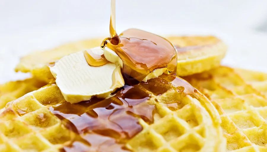 A close-up of a golden waffle with butter and maple syrup being poured on top, highlighting a tempting breakfast scene.