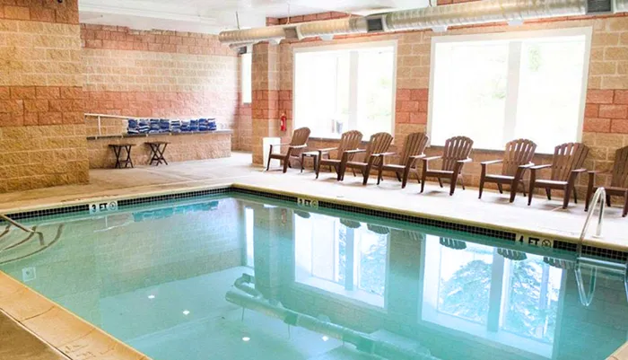 An indoor rectangular swimming pool is surrounded by brown chairs and tall windows set within a room with tan brick walls