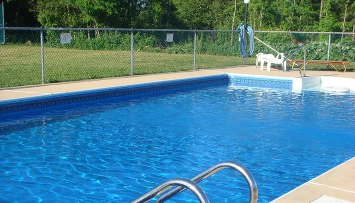 The image shows an outdoor swimming pool with clear blue water surrounded by a concrete deck a metal ladder on one side and a chain-link fence in the background on a sunny day