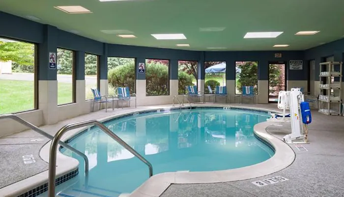 The image shows an indoor swimming pool with clear blue water surrounded by chairs and large windows letting in natural light