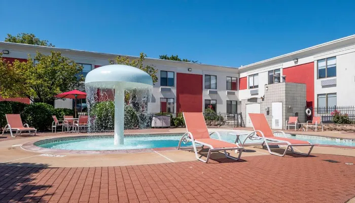A mushroom-shaped water feature is the centerpiece of this hotels outdoor swimming pool area surrounded by lounge chairs and flanked by a two-story building with a red and gray faade