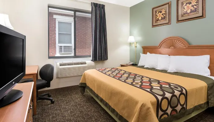 The image shows a neatly arranged hotel room with a double bed a desk with a chair a television and a window offering a view of a brick building