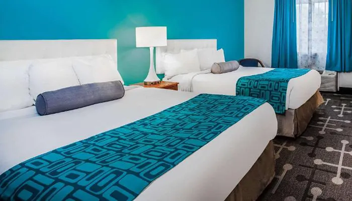 The image shows a brightly colored hotel room with two beds featuring white linens and contrasting blue decorative runners and pillows set against a vibrant blue accent wall