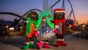 Three people in festive candy-themed costumes stand in front of a roller coaster at dusk.