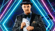 A person wearing a dark, patterned suit and a hat is holding a fanned deck of cards with a vibrant, geometric background.