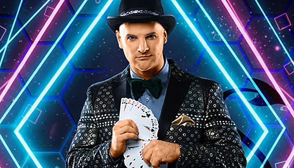 A person wearing a dark patterned suit and a hat is holding a fanned deck of cards with a vibrant geometric background