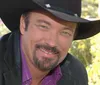 A man with a mustache is smiling at the camera wearing a black cowboy hat and a purple shirt under a black jacket with foliage and flowers in the background