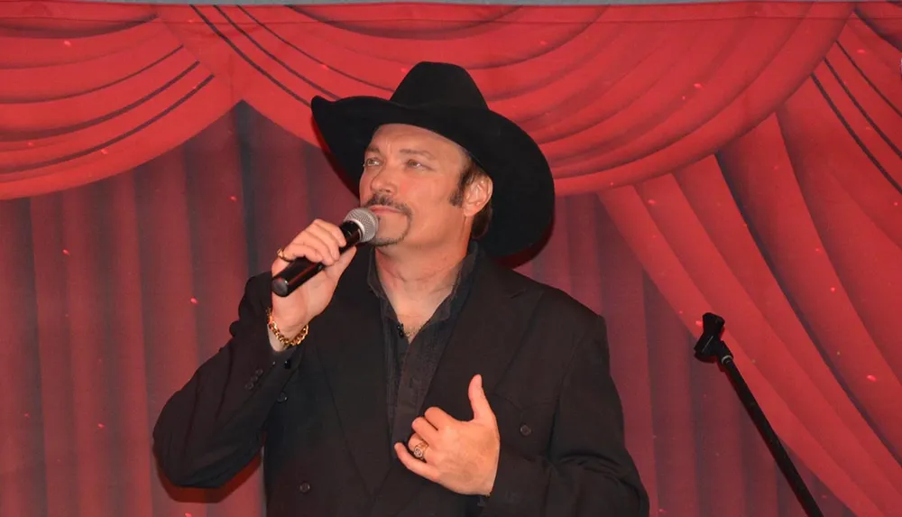 A man in a black cowboy hat is performing on stage with a microphone against a red draped background