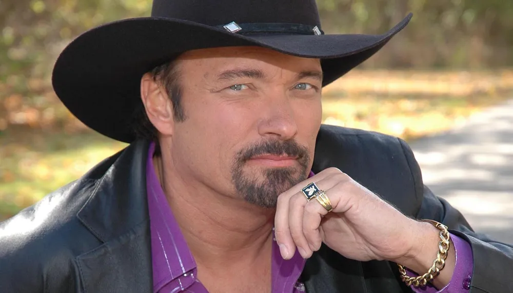 A man with a mustache wearing a black cowboy hat purple shirt and black jacket is posing with a thoughtful expression resting his chin on his hand adorned with a ring