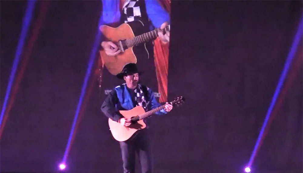 A musician in a cowboy hat plays an acoustic guitar on stage with a large projected image of himself playing in the background and colored stage lights shining in the foreground