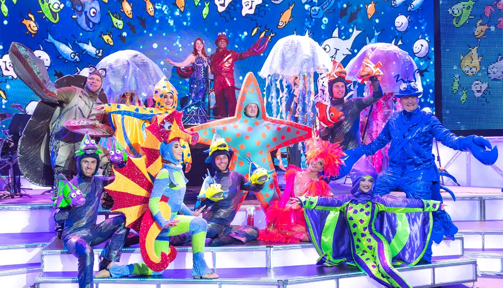 The image features a lively group of performers dressed in colorful elaborate costumes depicting sea creatures and marine-themed characters on a vibrant stage set