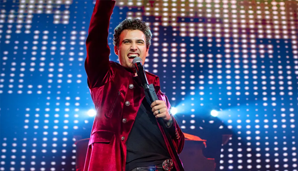 A performer in a red jacket is enthusiastically singing into a microphone with one arm raised against a backdrop of bright stage lights