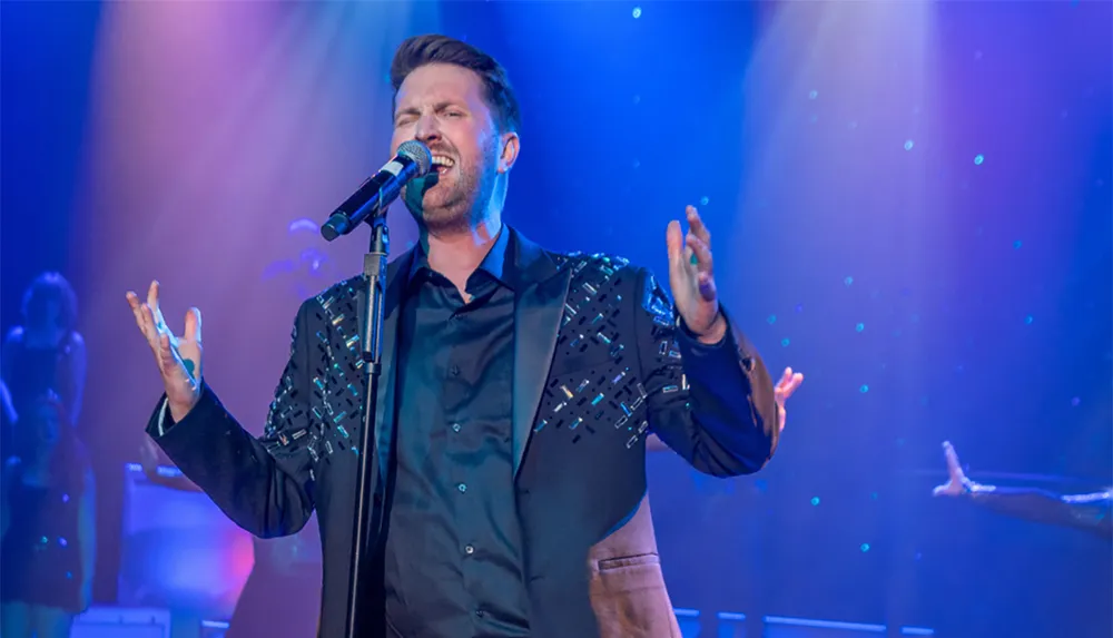 A man is passionately singing on stage with colorful lights illuminating him