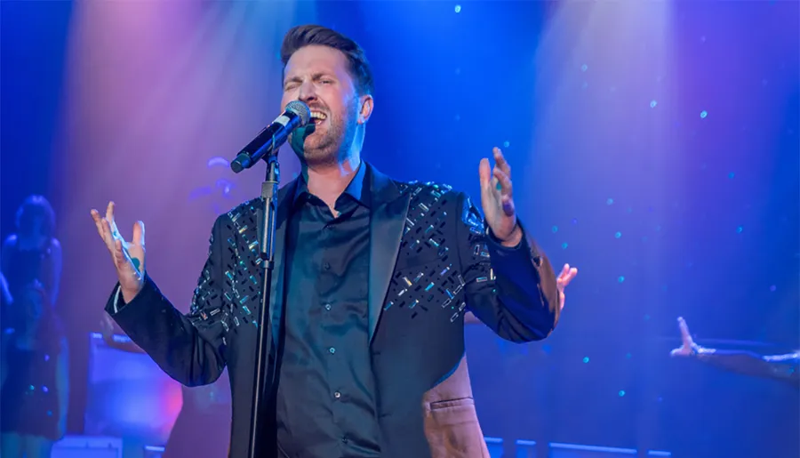 A man is passionately singing on stage with colorful lights illuminating him.