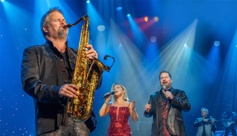 A group of musicians is performing on stage featuring a man playing the saxophone and two singers under colorful stage lights