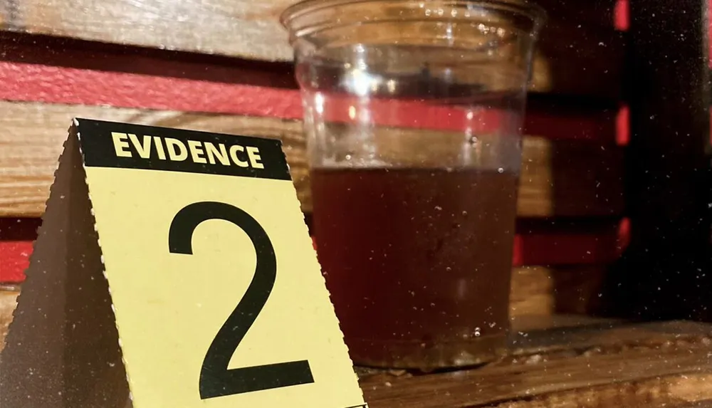 The image shows an EVIDENCE marker with the number 2 next to a half-filled glass of a dark liquid suggesting a scene from a criminal investigation