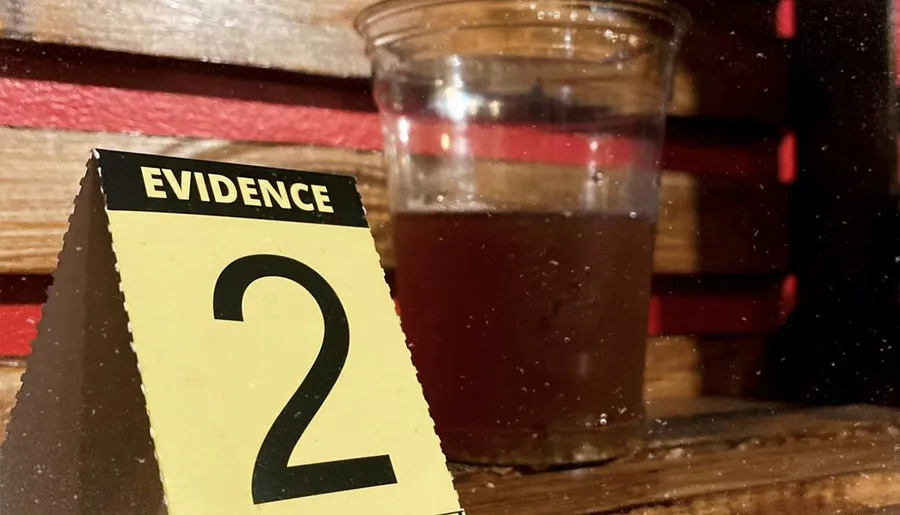 The image shows an EVIDENCE marker with the number 2 next to a half-filled glass of a dark liquid, suggesting a scene from a criminal investigation.