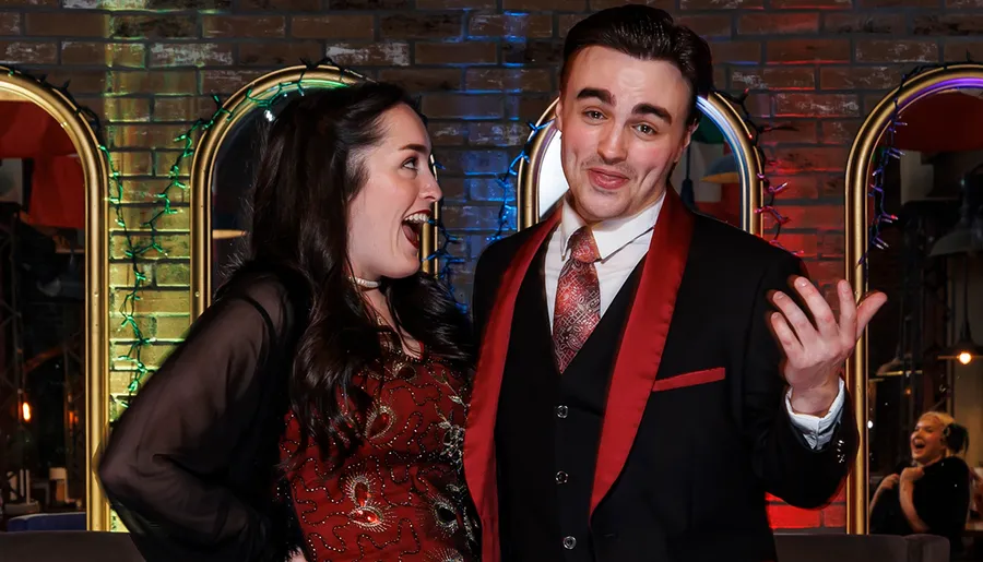 A woman and a man are happily posing, with both showing animated expressions against a backdrop of mirrors and colorful lights.