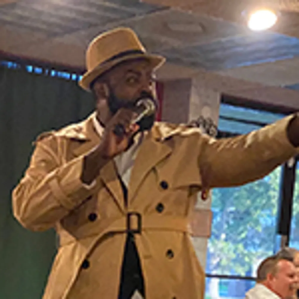 A man wearing a tan trench coat and hat is speaking into a microphone indoors