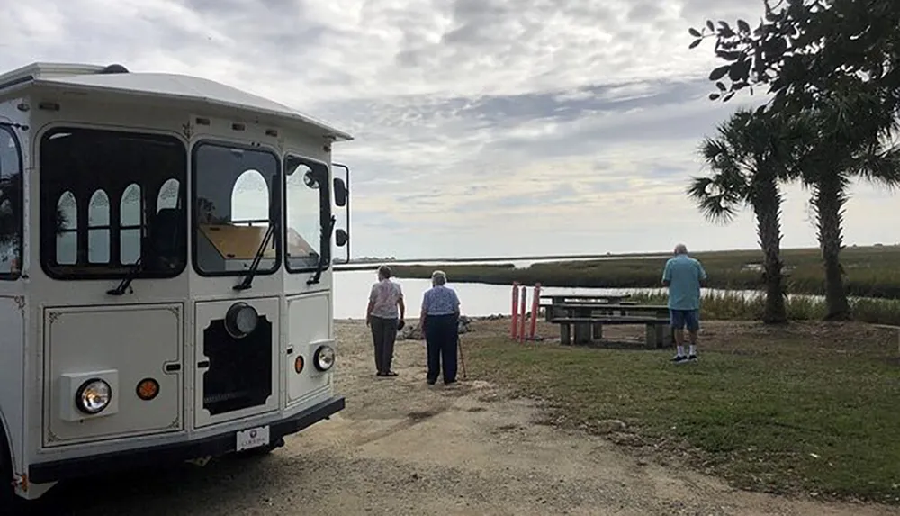 A trolley bus is parked near a scenic outdoor area where three individuals are walking towards a waterfront with a view of the marsh and a palm tree
