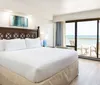 The image shows a neatly arranged hotel room with a large bed a picturesque ocean view through the balcony doors and a serene blue artwork above the bed