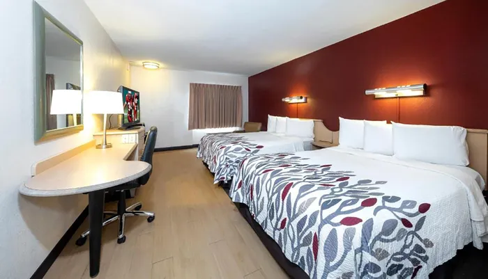 The image shows a neatly arranged hotel room with two double beds a work desk with a chair and a warm color scheme