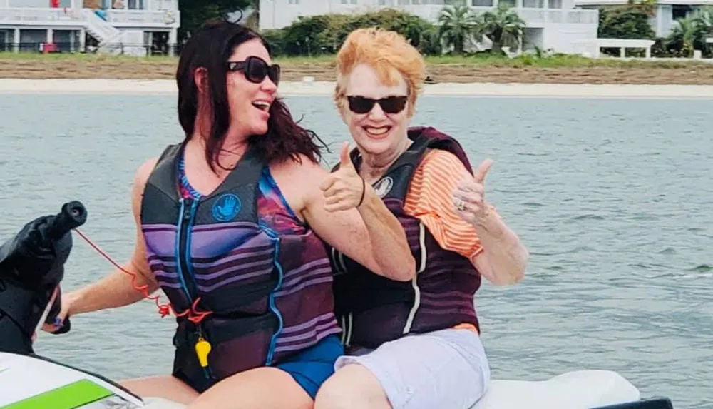 Two women are smiling and giving thumbs up while riding a jet ski on the water