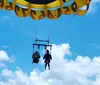 Two people are parasailing under a smiling face-decorated parachute against a clear blue sky