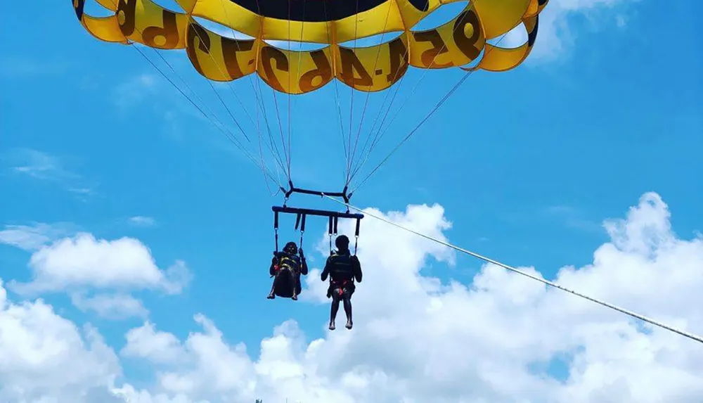 Two people are parasailing under a yellow and black canopy suspended high in the sky on a sunny day