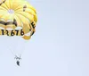 Two people are parasailing under a smiling face-decorated parachute against a clear blue sky