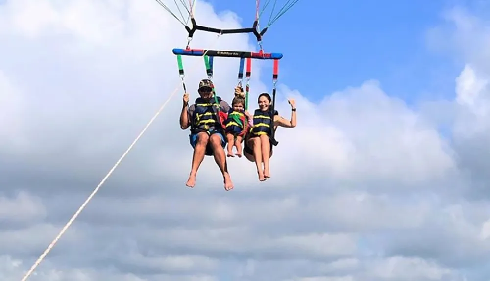 Three individuals are parasailing high above the ground with clear blue skies and fluffy white clouds in the background