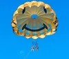 Two people are parasailing under a smiling face-decorated parachute against a clear blue sky