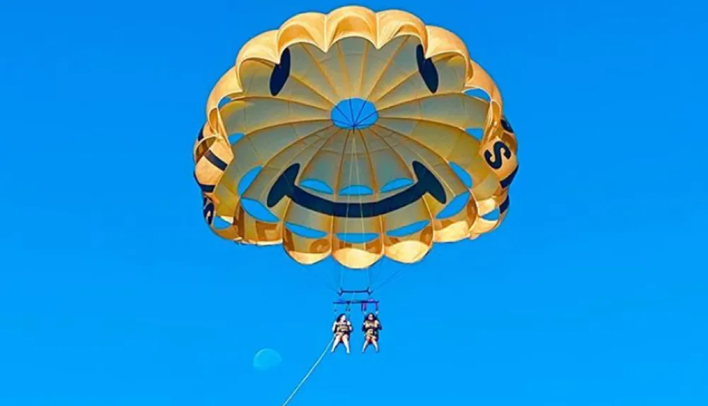 Two people are parasailing under a smiling face-decorated parachute against a clear blue sky