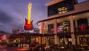 This is an evening photo of the Hard Rock Cafe, with its iconic neon guitar sign, situated in a lively outdoor shopping and dining area.