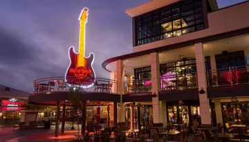 This is an evening photo of the Hard Rock Cafe, with its iconic neon guitar sign, situated in a lively outdoor shopping and dining area.