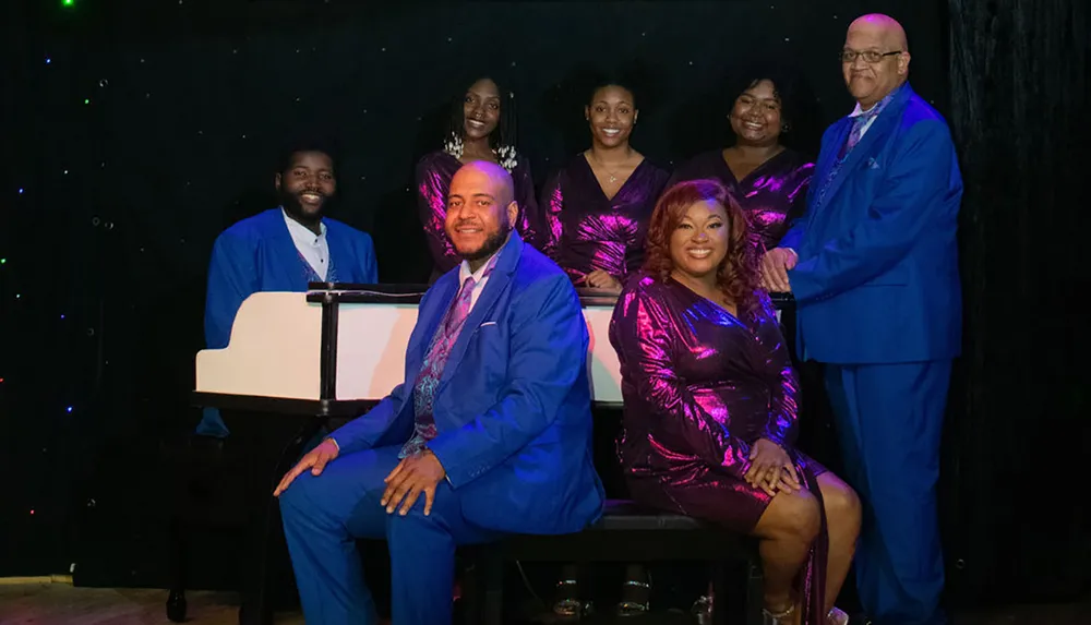 A group of seven elegantly dressed people four women and three men are posing for a photo seemingly in a celebratory or performance setting with a starry background and a piano