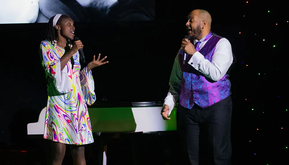Two people are standing on a stage holding microphones seemingly engaged in a duet or a conversation in a performance setting with colorful lights in the background