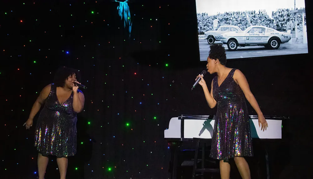 Two performers in sequined dresses are singing on a stage against a backdrop with a vintage car photo and twinkling colored lights