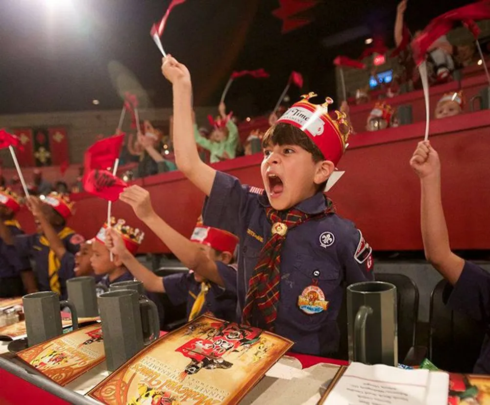 A young boy in a scout uniform is enthusiastically waving a red flag while wearing a crown sitting at a table with menus and other children in a festive atmosphere