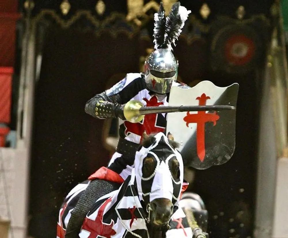 A person in medieval knight armor is mounted on a decorated horse holding a jousting lance and a shield while charging forward likely at a reenactment event or a tournament show