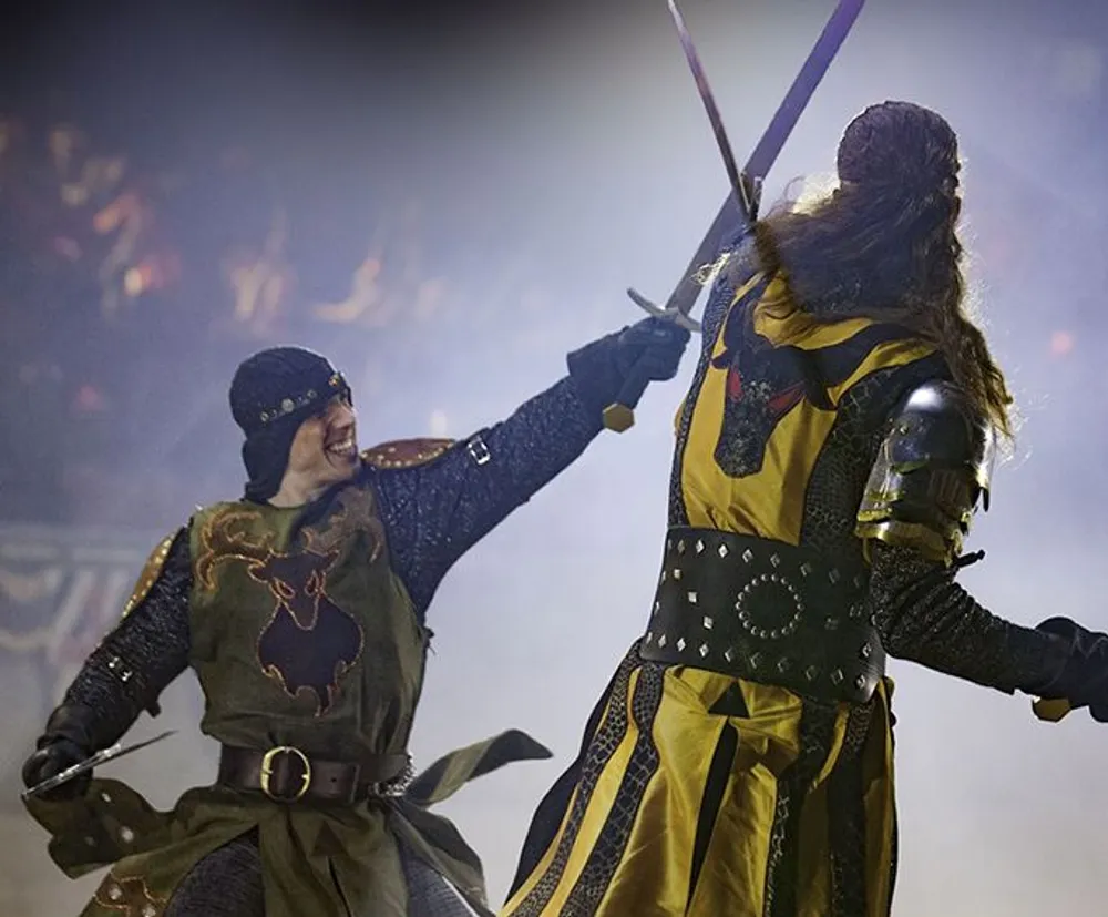 Two individuals in medieval-style costumes are engaged in a staged sword fight before an audience