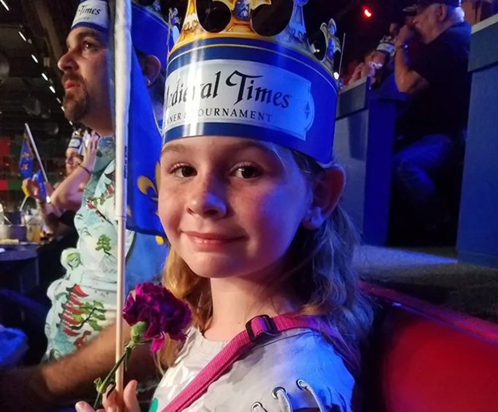 A smiling young girl wearing a paper crown and holding a rose is at what appears to be a Medieval Times dinner and tournament experience with spectators and flags in the background