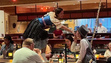 A woman in a festive costume stands on a table and playfully interacts with a person wearing a top hat in a busy restaurant setting.