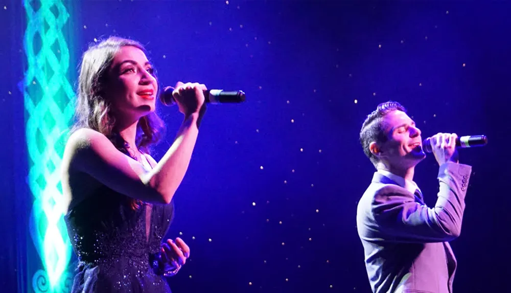 Two vocalists are performing on stage under blue lighting with the woman in focus at the forefront and the man slightly out of focus in the background
