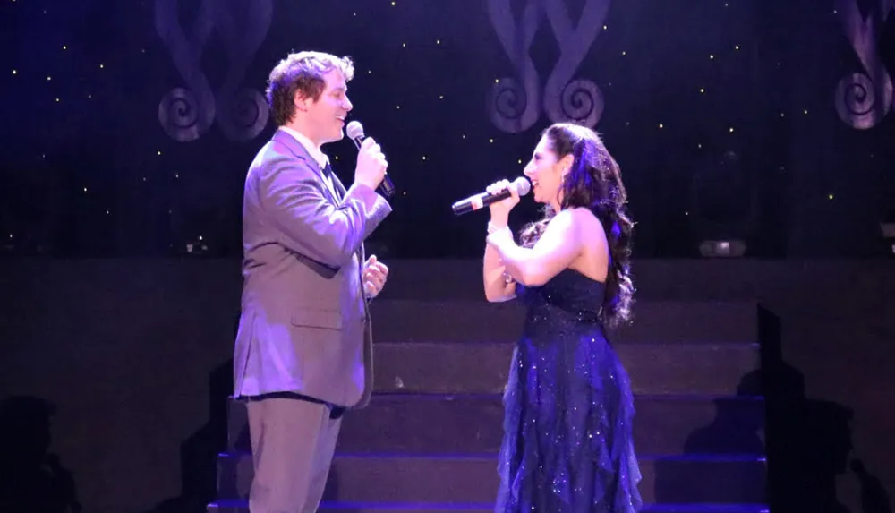 Two singers are performing a duet on stage with the man wearing a suit and the woman in a blue gown both holding microphones