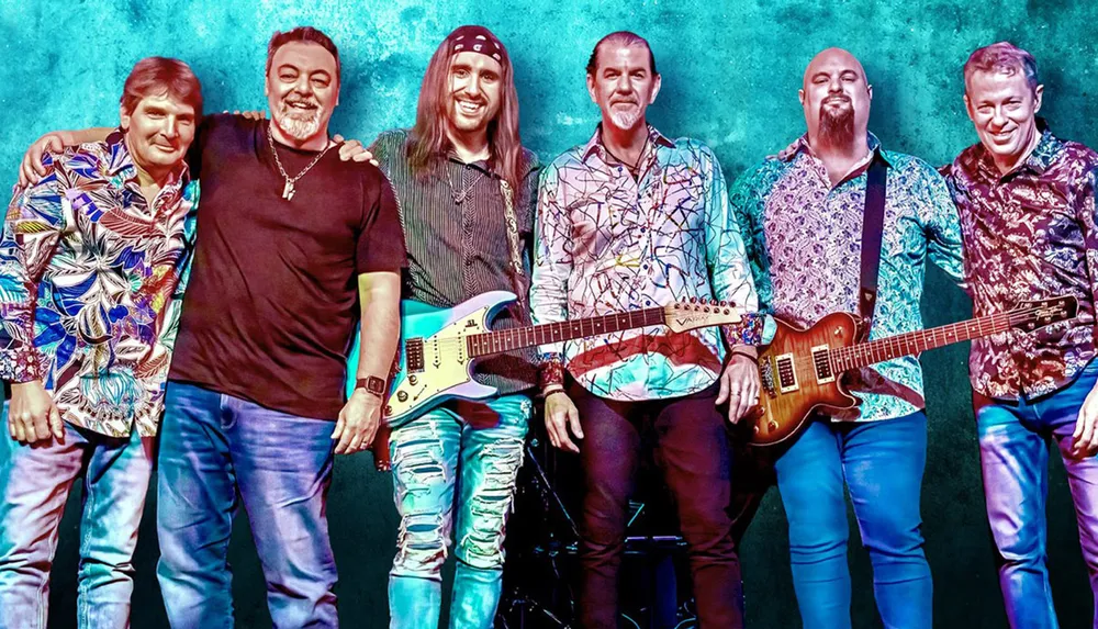 Six smiling men likely a band are posing together with two of them holding electric guitars against a vibrant teal background lit with stage lighting effects