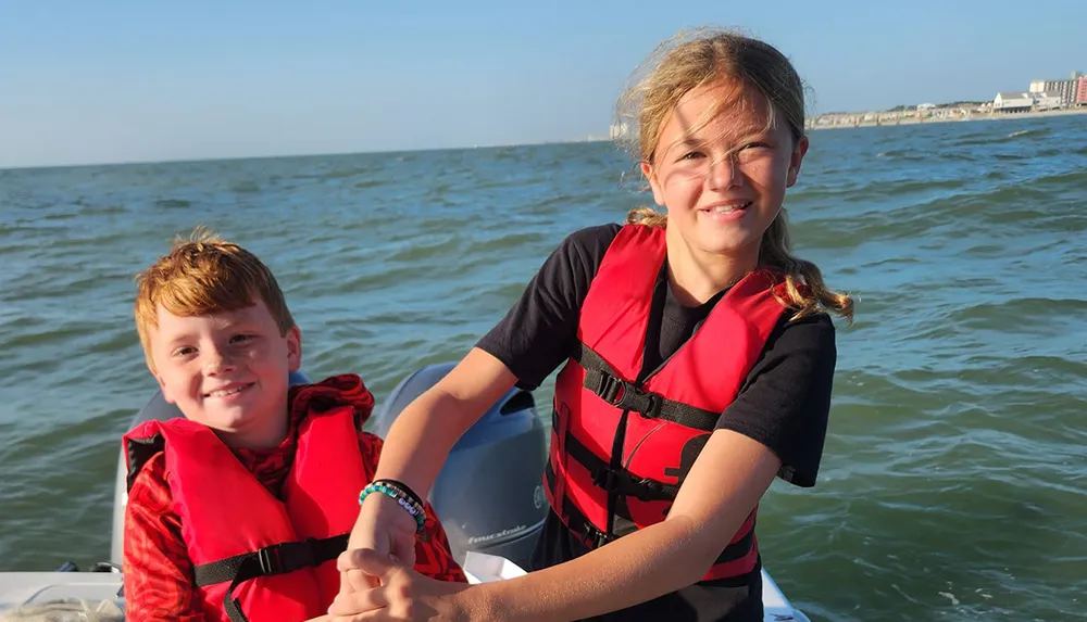 Two smiling children wearing red life jackets are seated on a boat with the ocean in the background